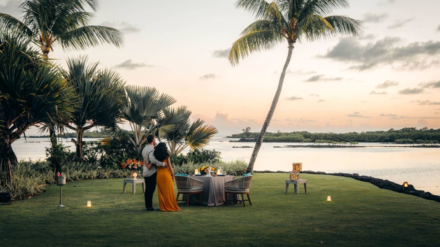 A couple stand on the shore, arms around each other, gazing toward the ocean; set table with two chairs beside swaying palms.