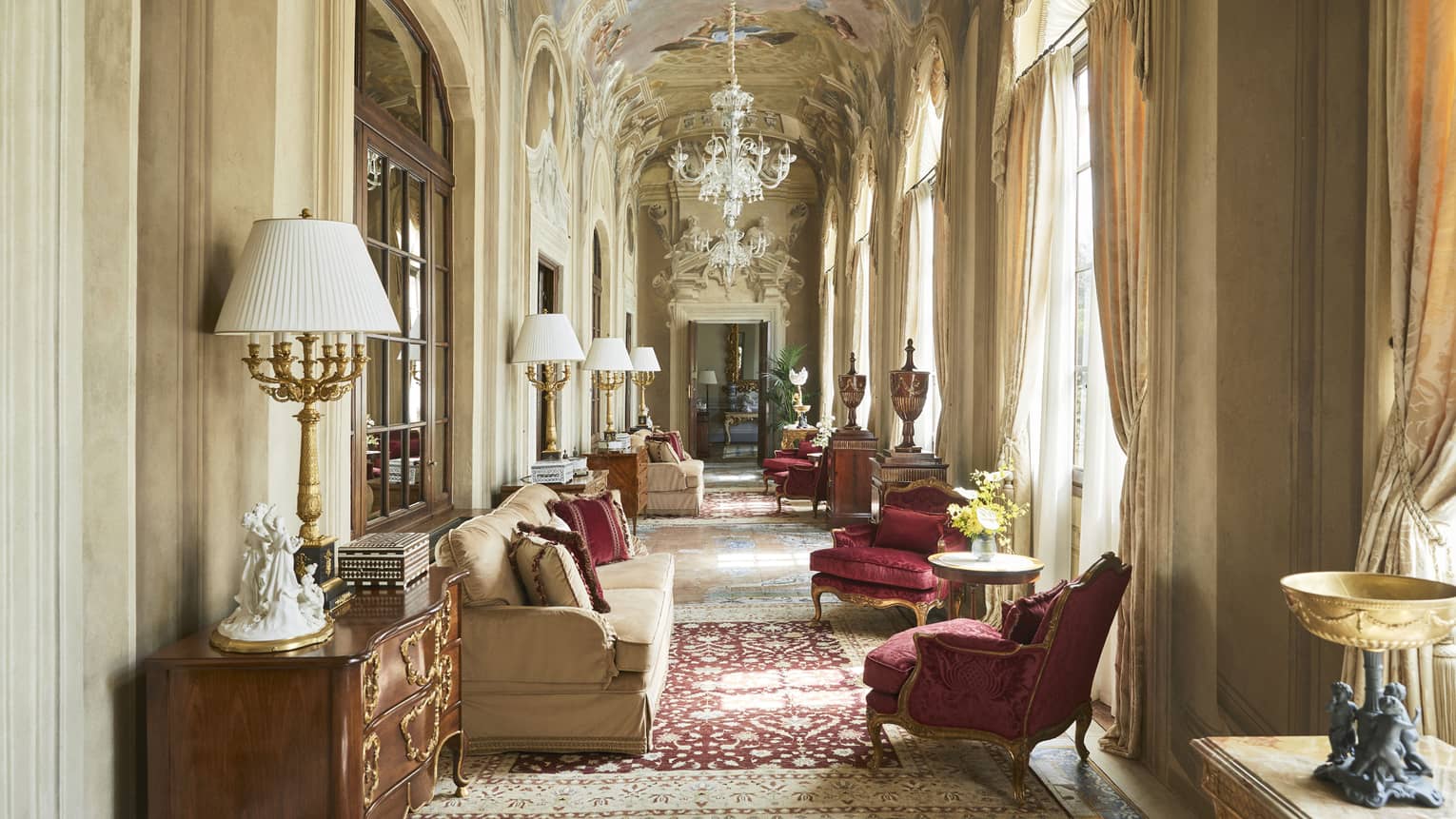 Grand, arched hallway with frescoed ceiling and chandelier, lounge seating and ruby-red arm chairs, in the Royal Suite at Four Seasons Hotel Florence