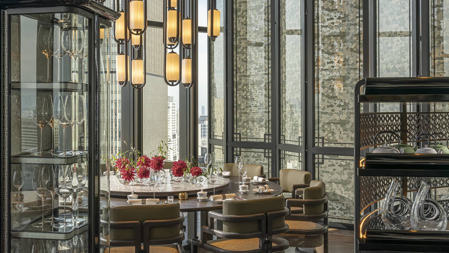 A mod chandelier hangs over red chrysanthemums on a round dinner table set beside tall windows and a vitrine of wine glasses.