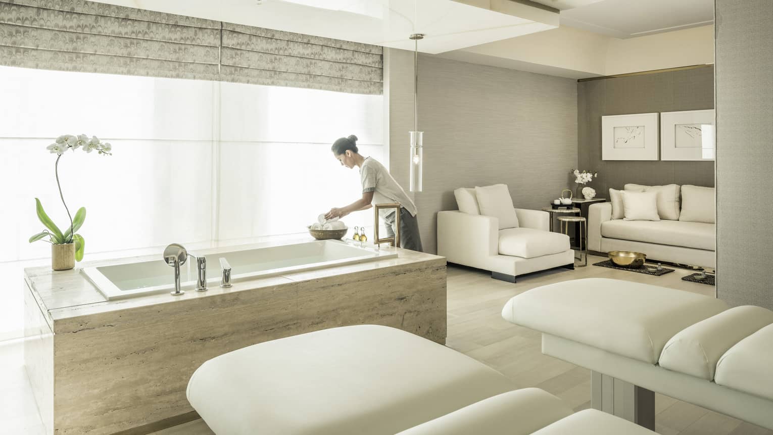 A spa attendant arranges some products in the spa suite, with two massage beds and a soaking tub in front