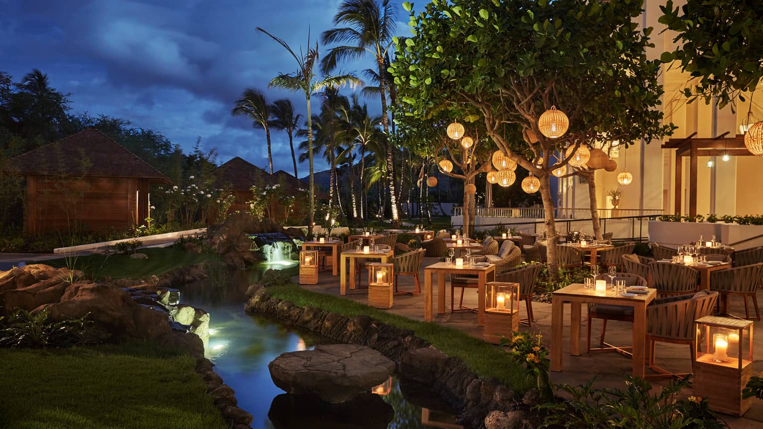 Noe patio dining tables, chairs, lanterns by stream at night