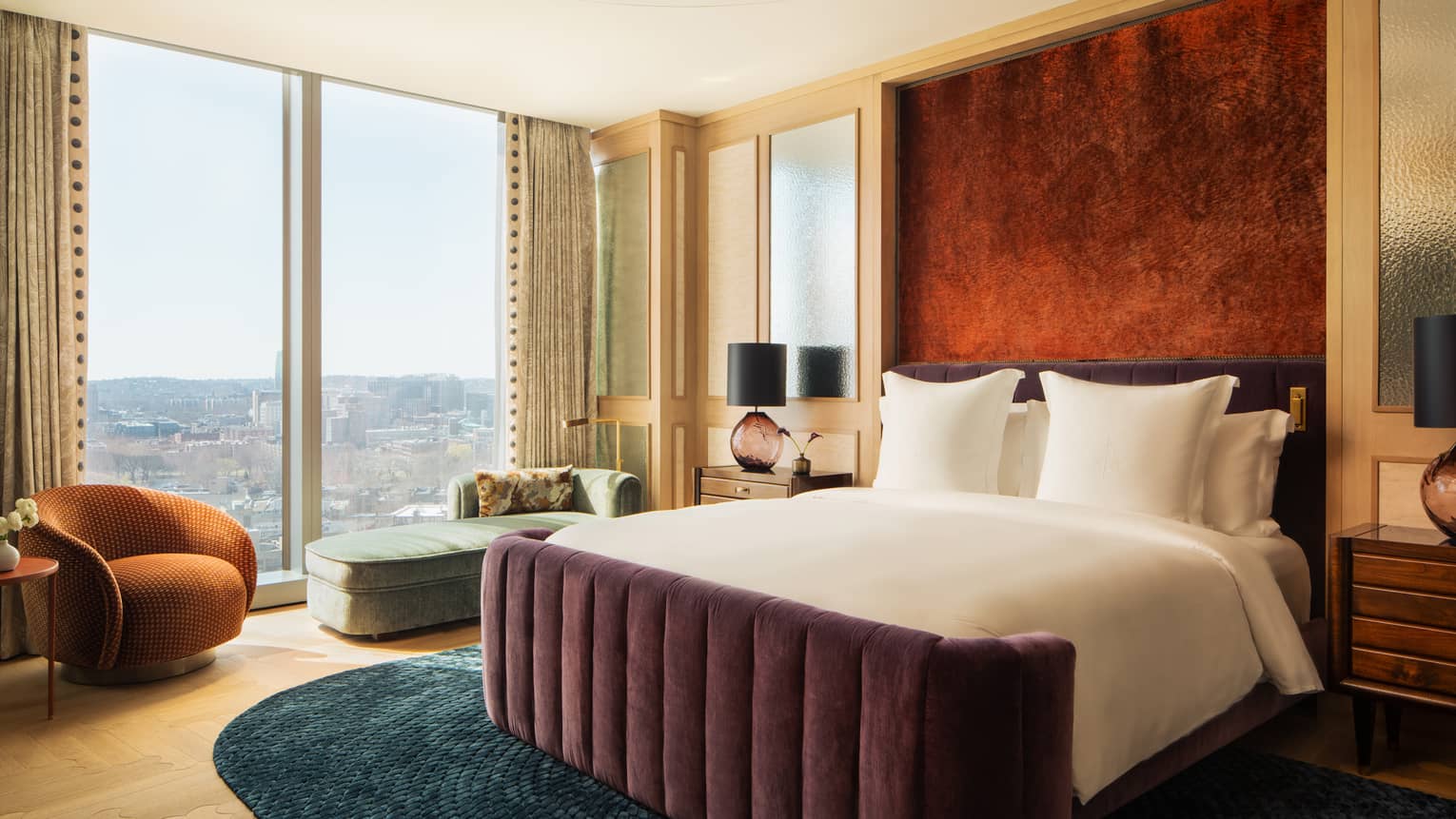 Contemporary bedroom with a king-sized velvet bed frame, wall of windows with city views, chaise and arm chair