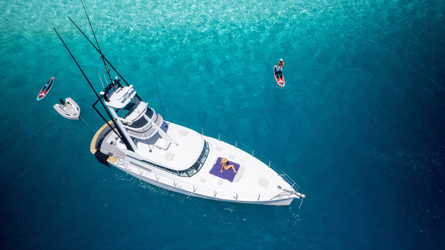 Aerial shot of yacht with person lying on deck, several smaller boats surrounding it in turquoise water