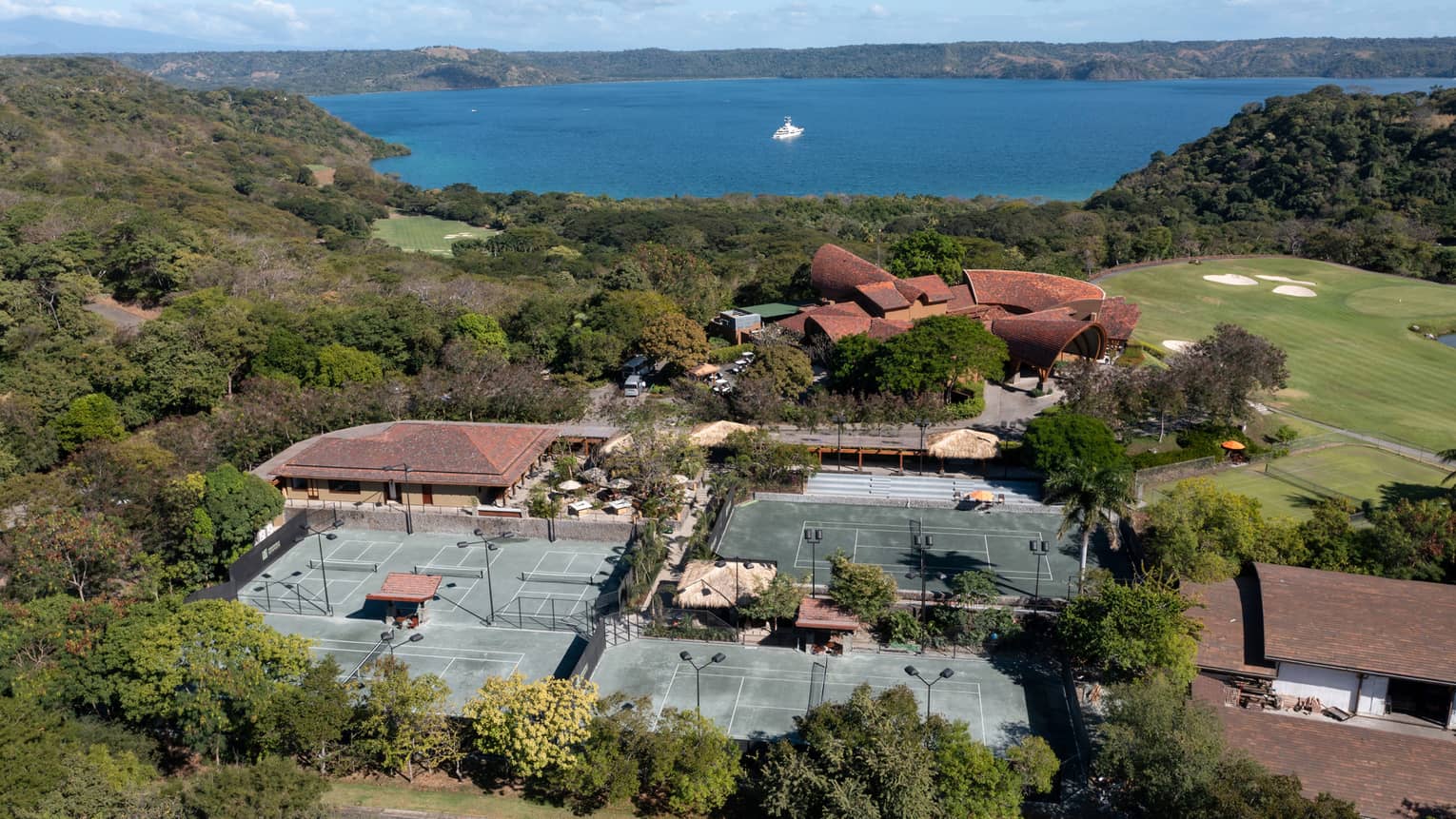 Aerial view of resort tennis centre, surrounded by trees with the ocean in the near distance
