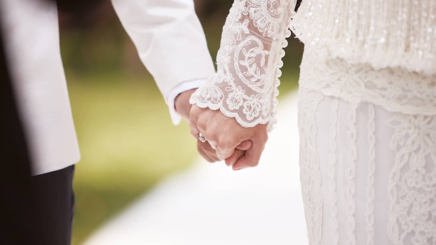 Close-up of bride with lace sleeve and groom with white suit holding hands 