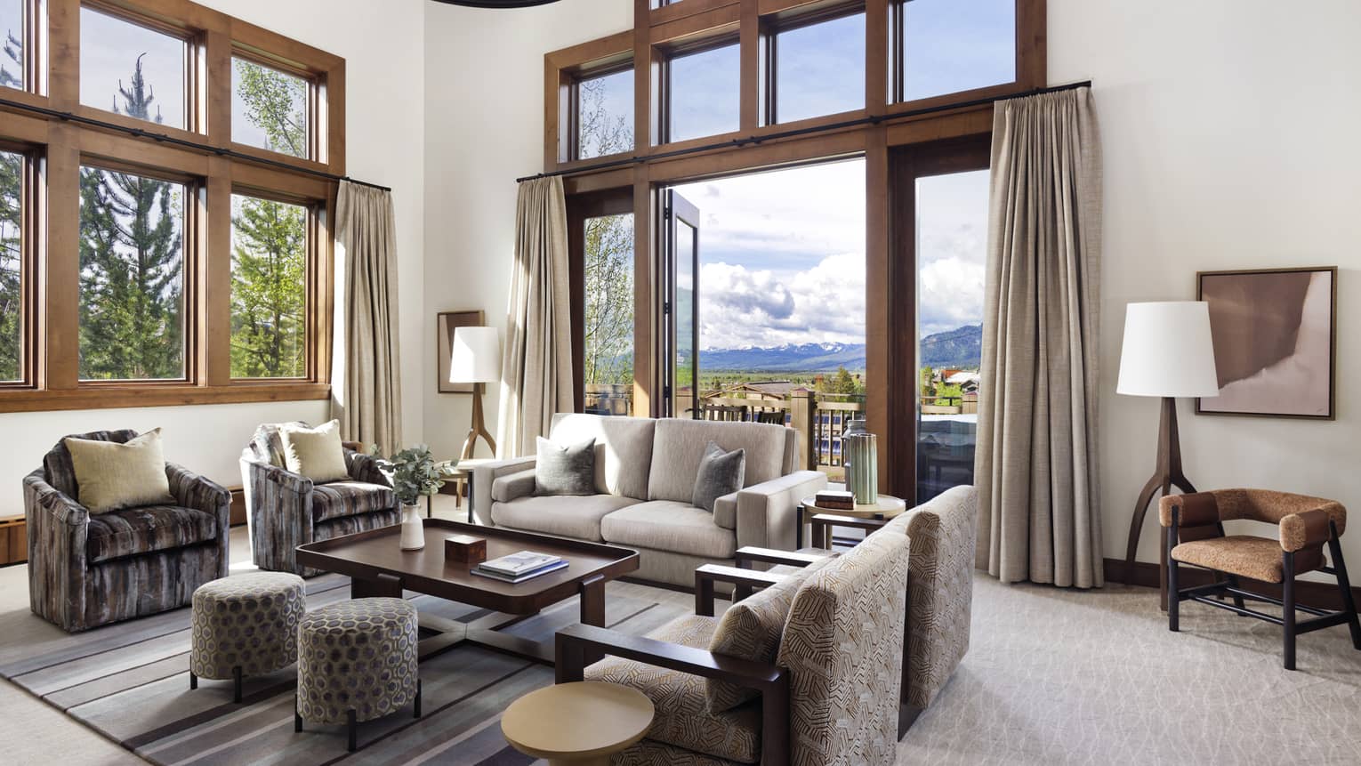 Luxury resort living room with high ceilings, large windows, sofa, armchairs, ottomans, table, floor lamps and doors opening to a mountain-view balcony