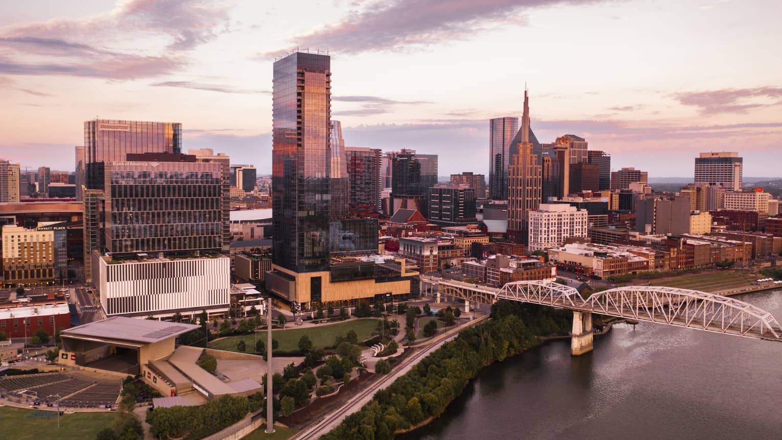 Aerial view of downtown Nashville, Tennessee at sunset, featuring modern skyscrapers, a river, a bridge, and a park with greenery in the foreground.