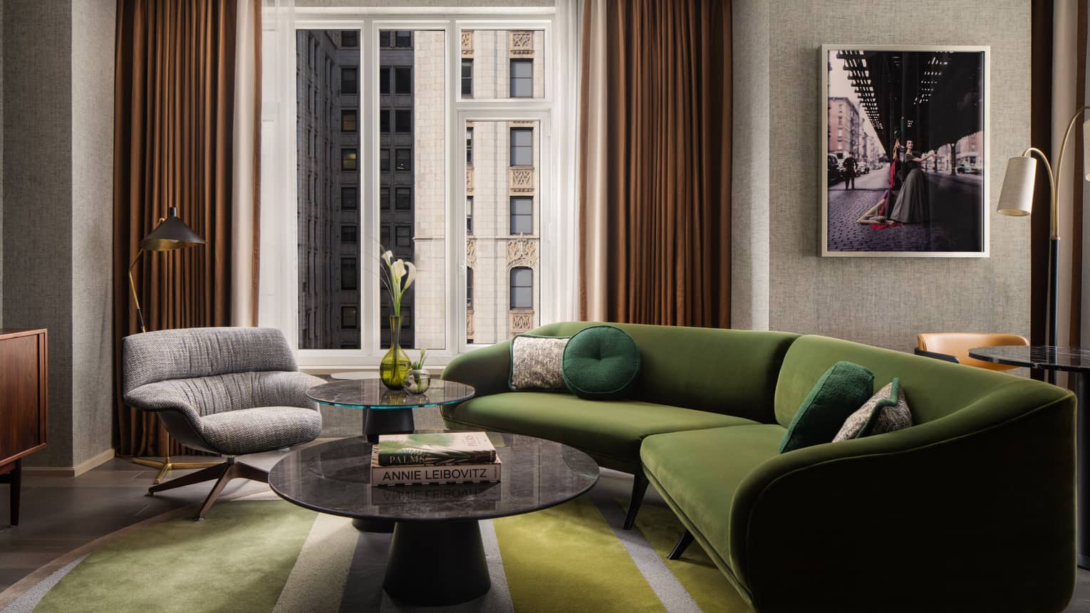 Suite living room with green sectional sofa, round coffee tables, gray armchair, wall art, and large window overlooking city buildings