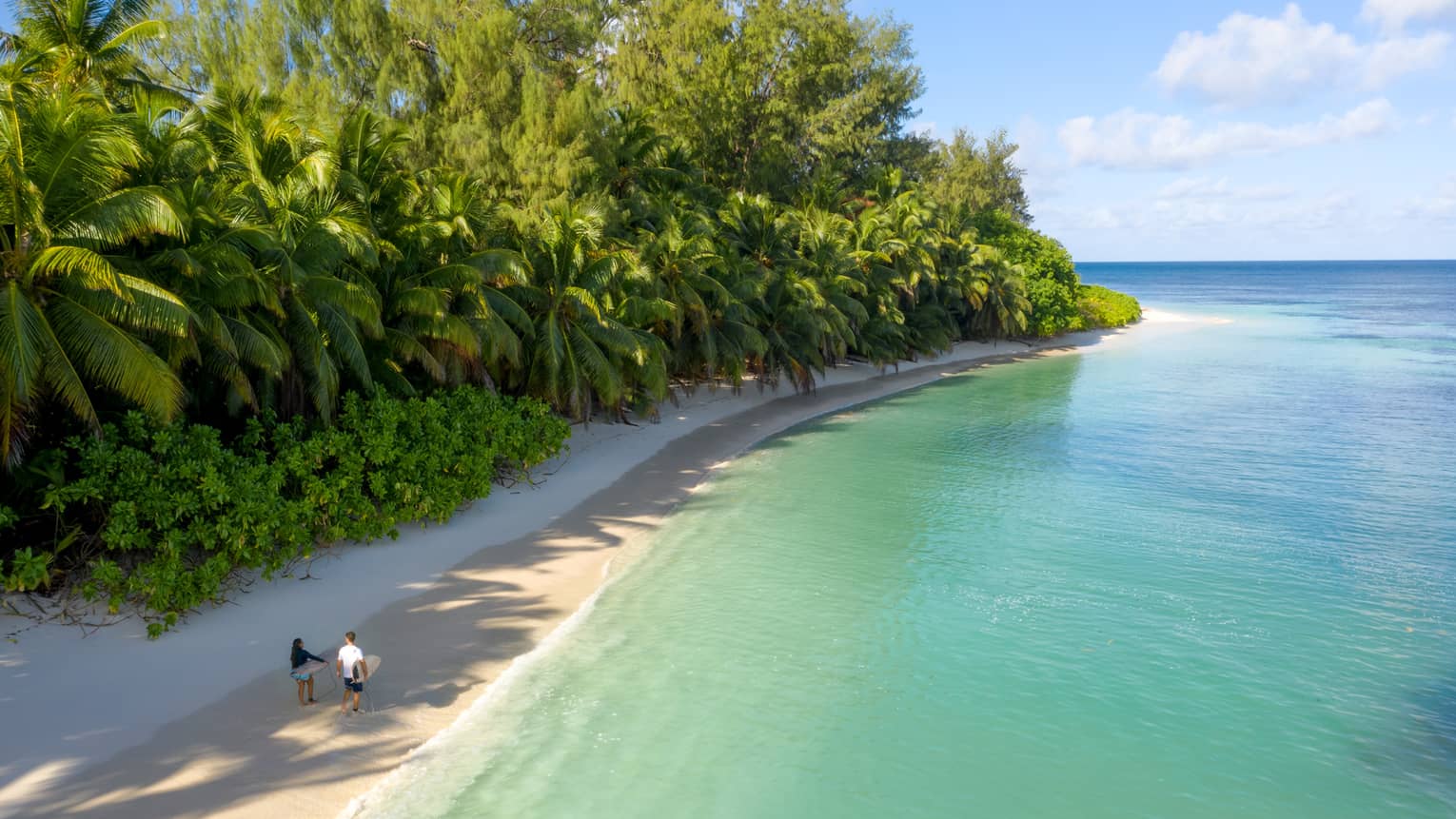 Couple walking along beach, carrying surfboards, flanked by lush tropical vegetation and blue-green water