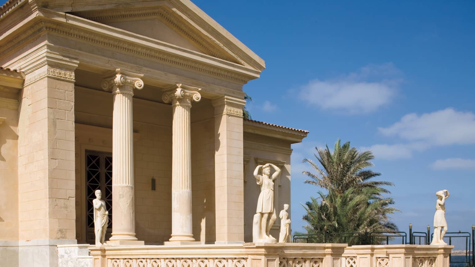 Stone building with columns, statues and palm tree against blue sky background