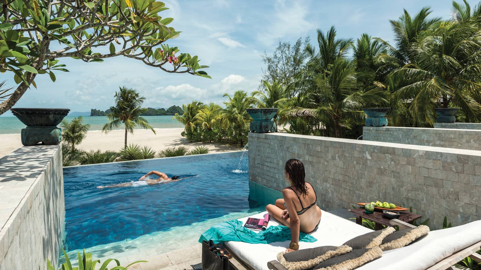 A woman sits on a lounge chair by an infinity pool built on the edge of the beach. 