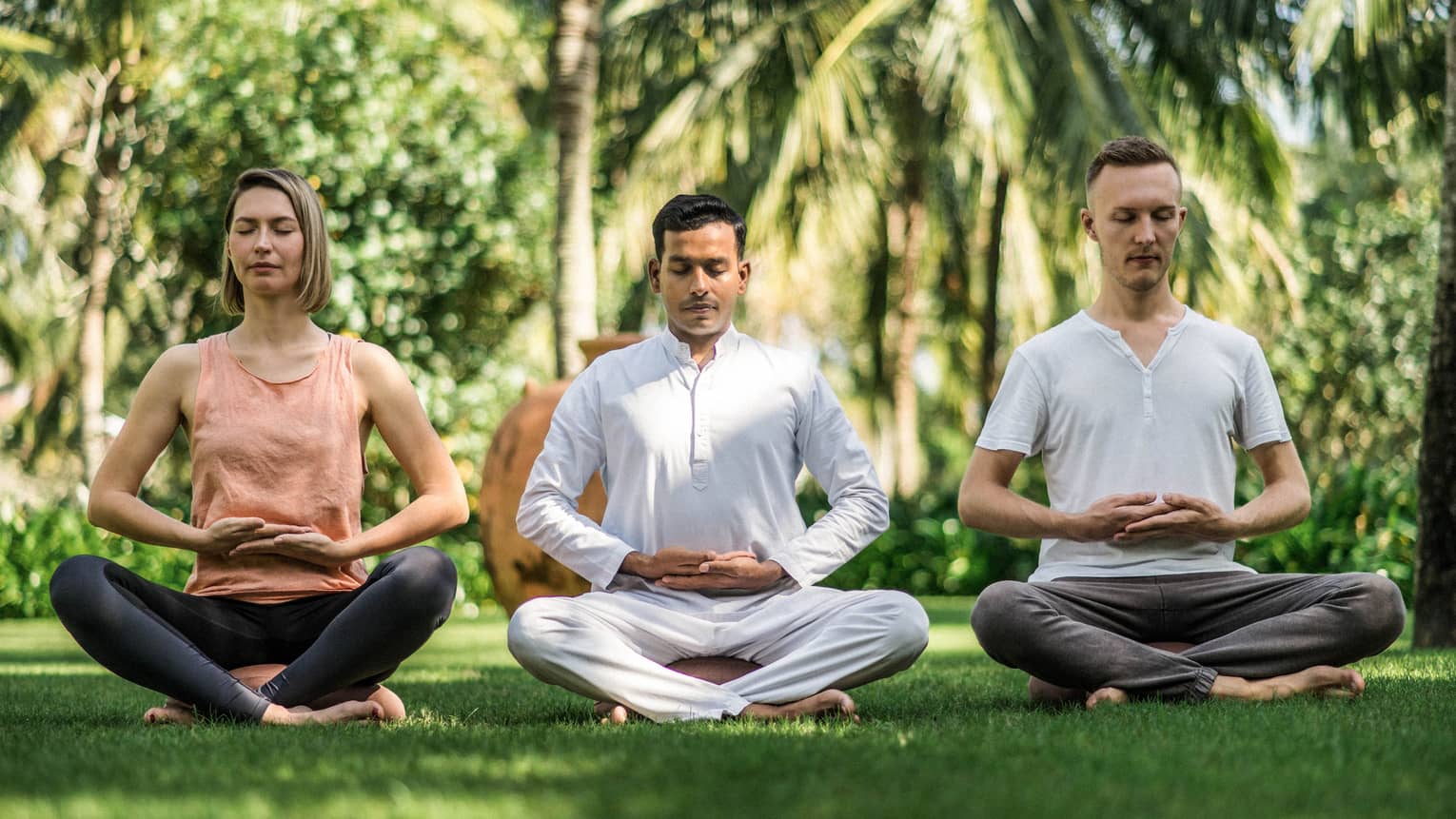 Three people meditating in a seated position on a grassy lawn, surrounded by tropical greenery, with their hands resting gently on their laps.