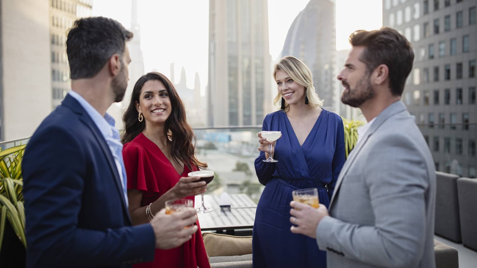 Four people dressed in cocktail attire stand talking and holding drinks on a rooftop terrace with a city skyline in the background