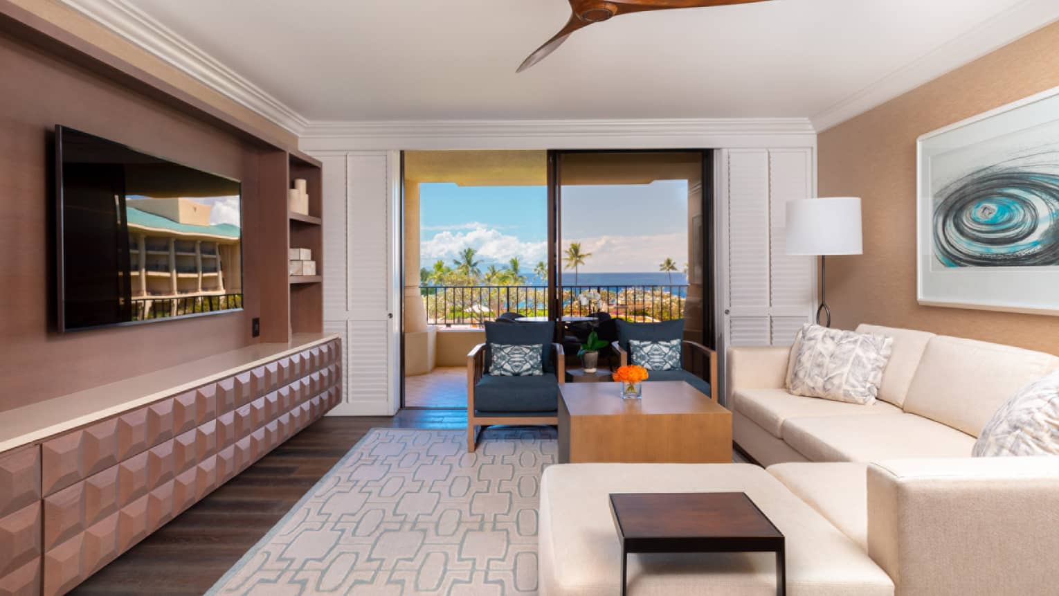 Living room with beige sofa, wall-mounted TV, ceiling fan and sliding doors opening to a balcony with ocean view
