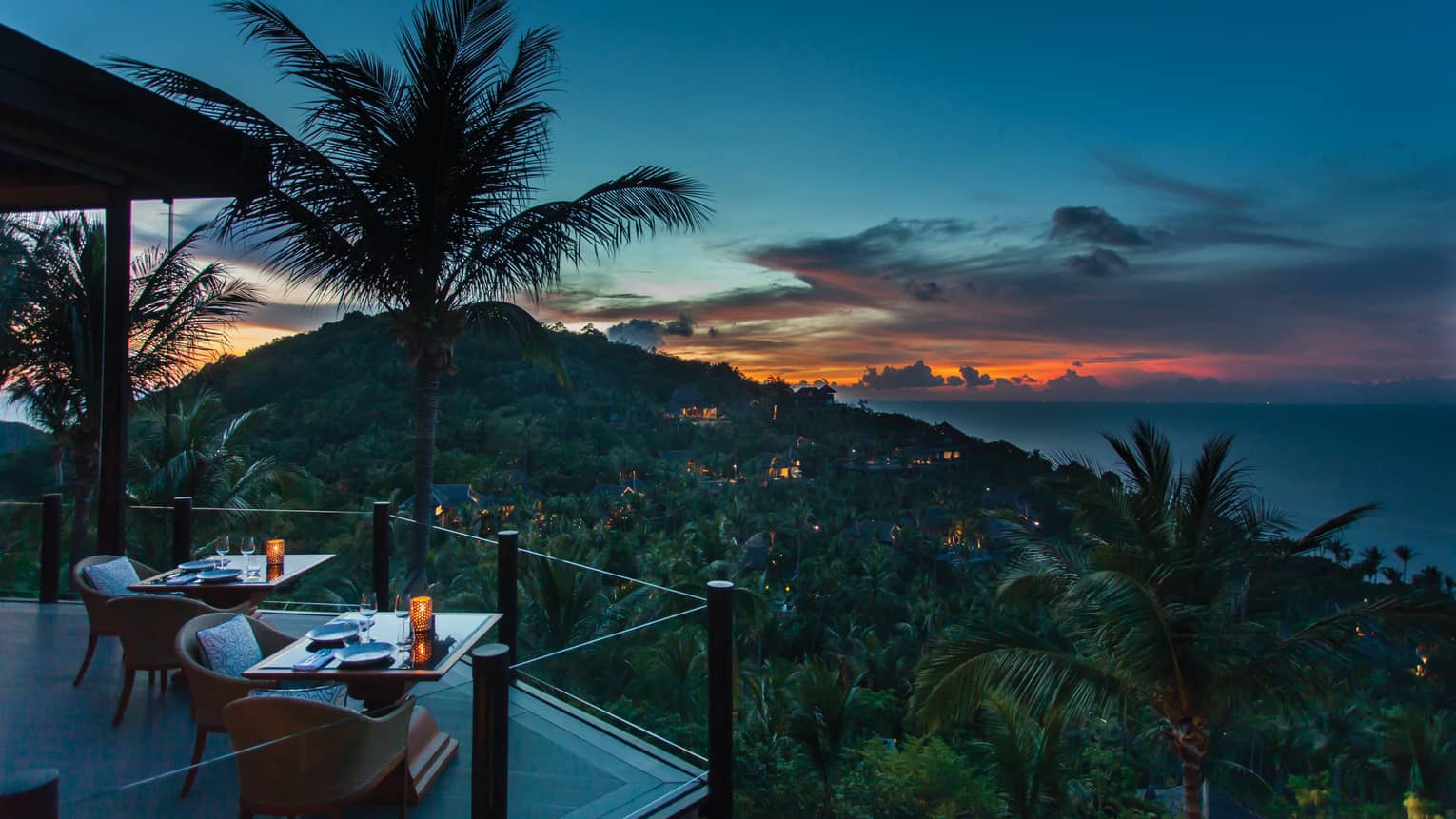 Corner of extended balcony with dining tables and chairs at dusk, expanse of forest, palm trees