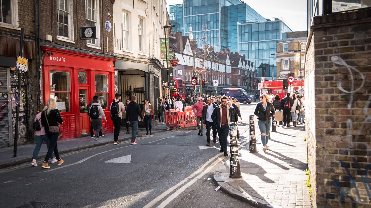 Crowds stroll along sunny East End street in London, past brick buildings, shops