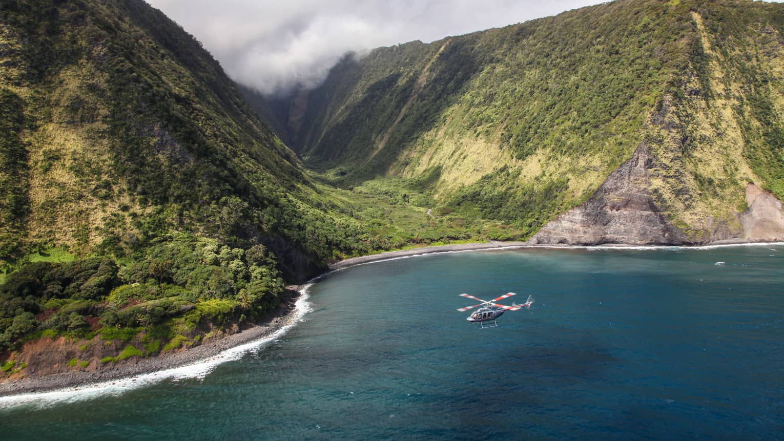 Helicopter flies over ocean by sweeping green volcanic mountains