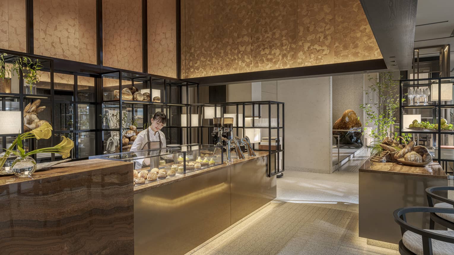 A person stands behind a counter displaying food items in an open, well-lit space decorated in warm neutral tones; the space also includes chairs, open shelves, an additional counter and a freestanding island