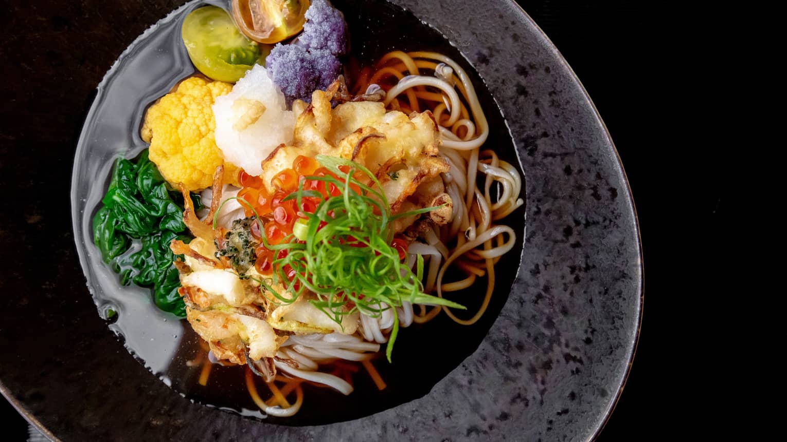 A close-up of a noodle dish in a broth with colourful vegetables, deep-fried meat and fish eggs in a dark grey bowl.