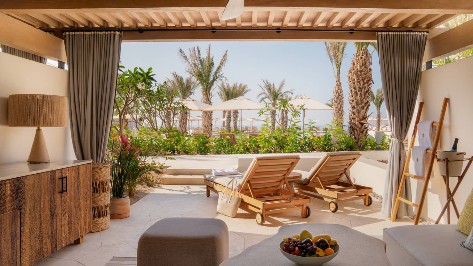 View from inside a neutral cabana with champagne, a fruit bowl and lounge chairs facing out towards palm trees and umbrellas.