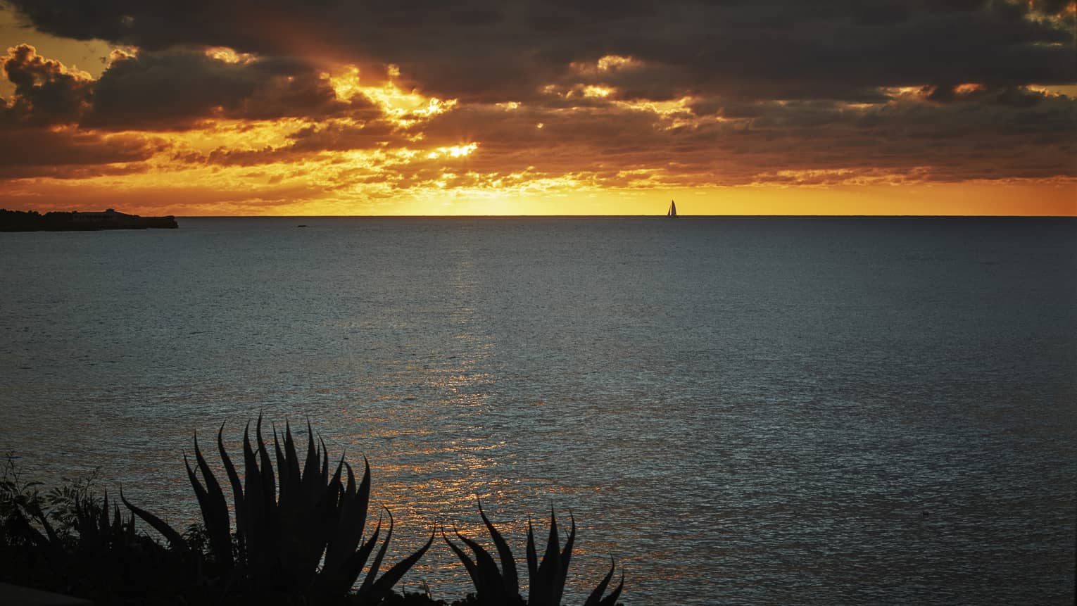 Bright orange sunset over the ocean with dramatic clouds and a sailboat in the distance