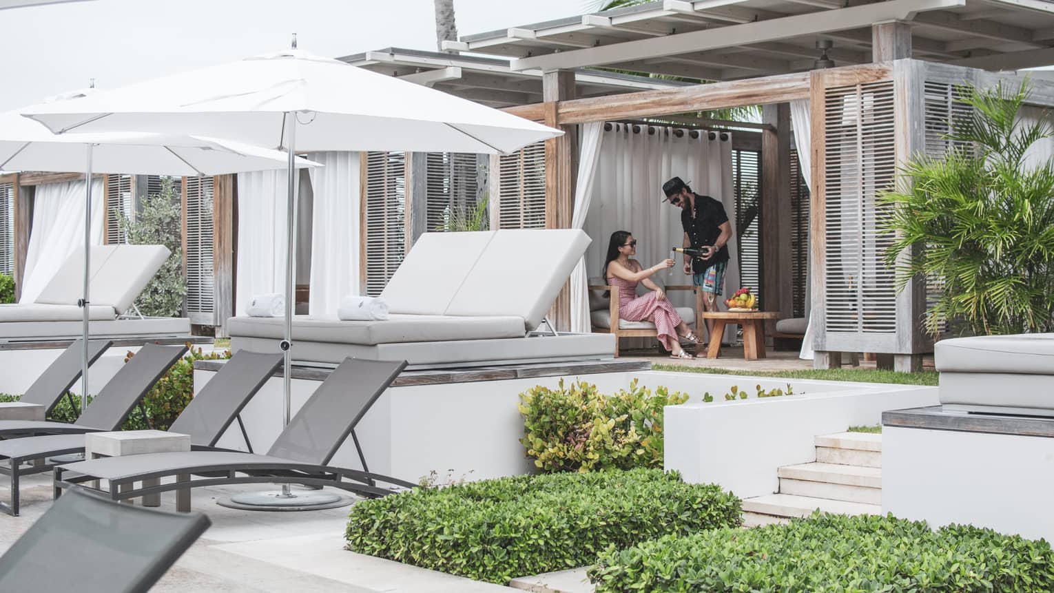 Two talk to each other in a covered pool cabana with a row of white daybeds and a row of grey lounge chairs and white umbrellas in front of them