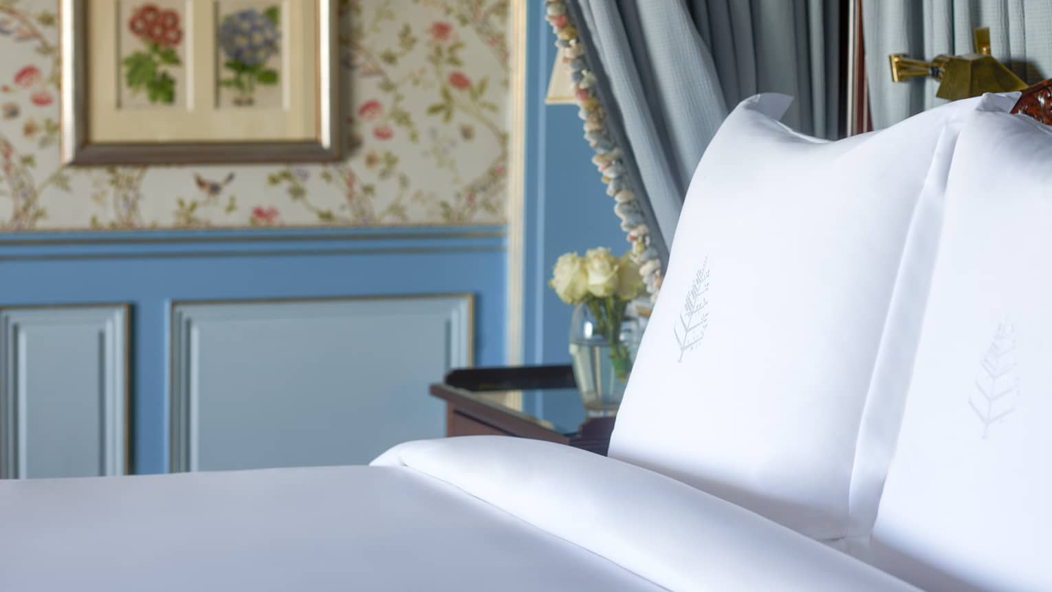 Close-up of bed with white linens, side table with flowers, blue curtains, paisley wallpaper