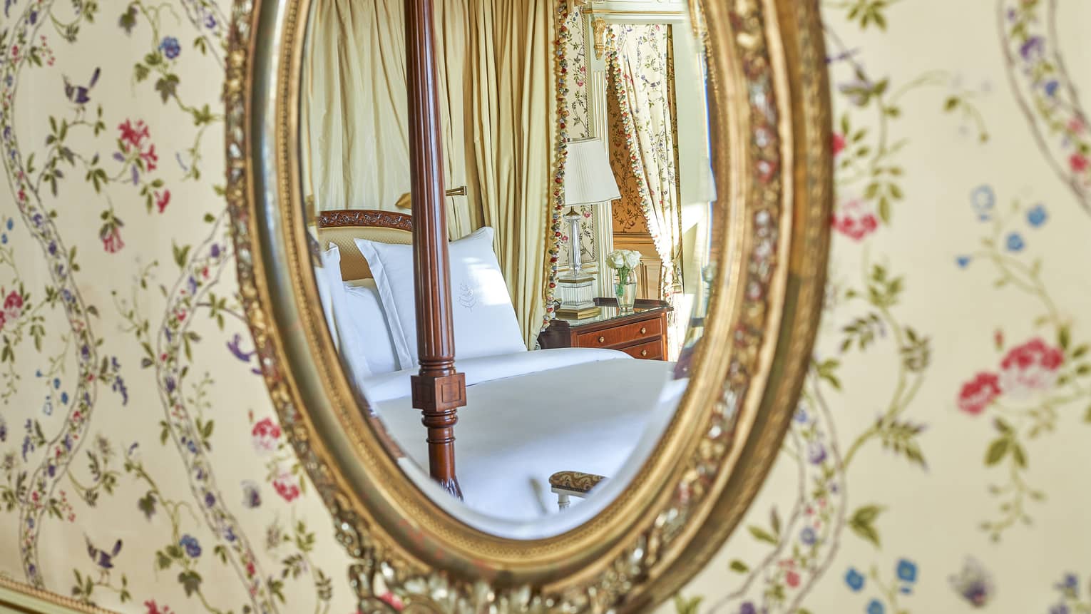 Close-up of oval gold-framed mirror and paisley wallpaper with reflection of bed