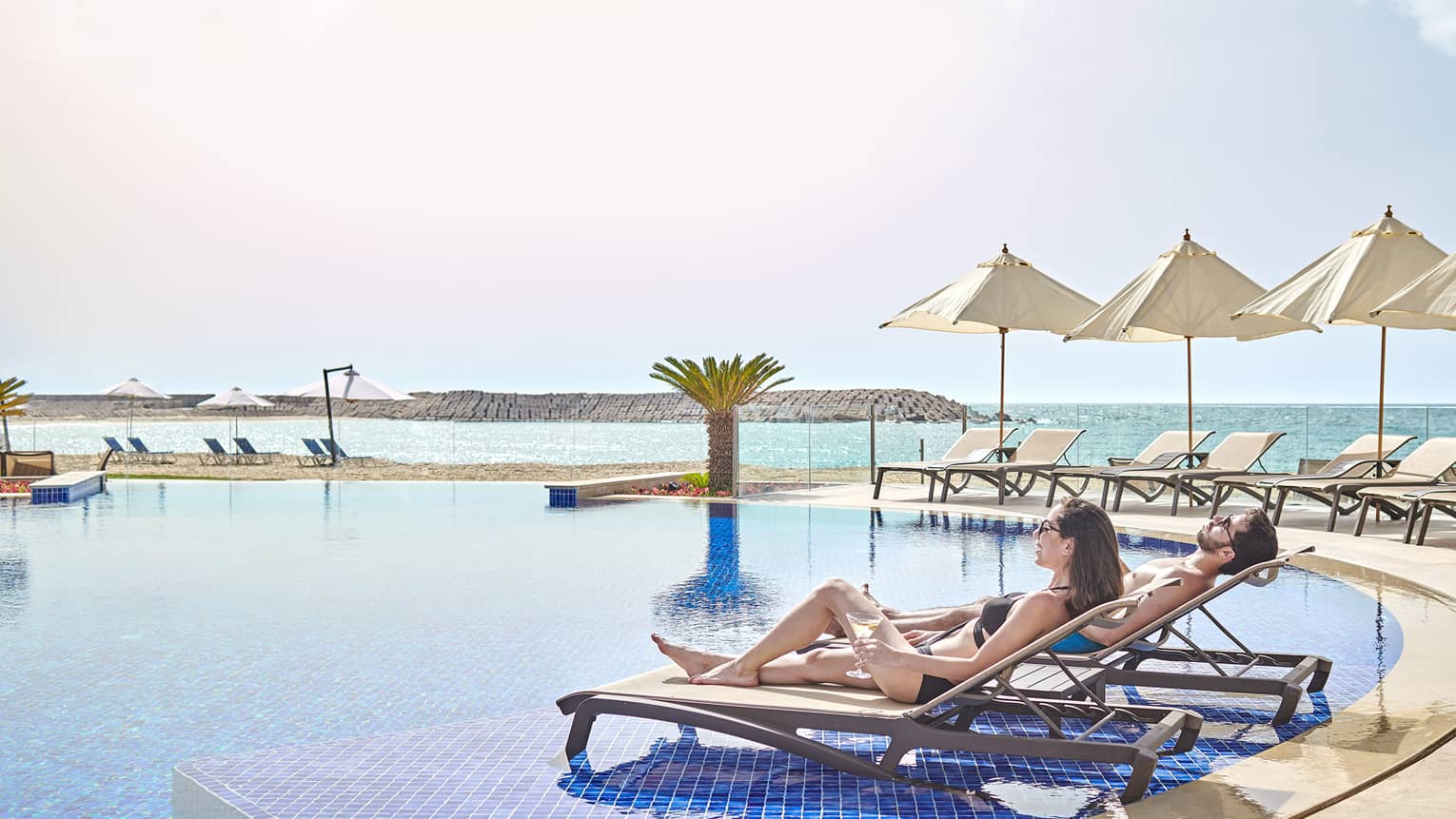 A couple lounges by the pool with view of ocean, beach umbrellas 