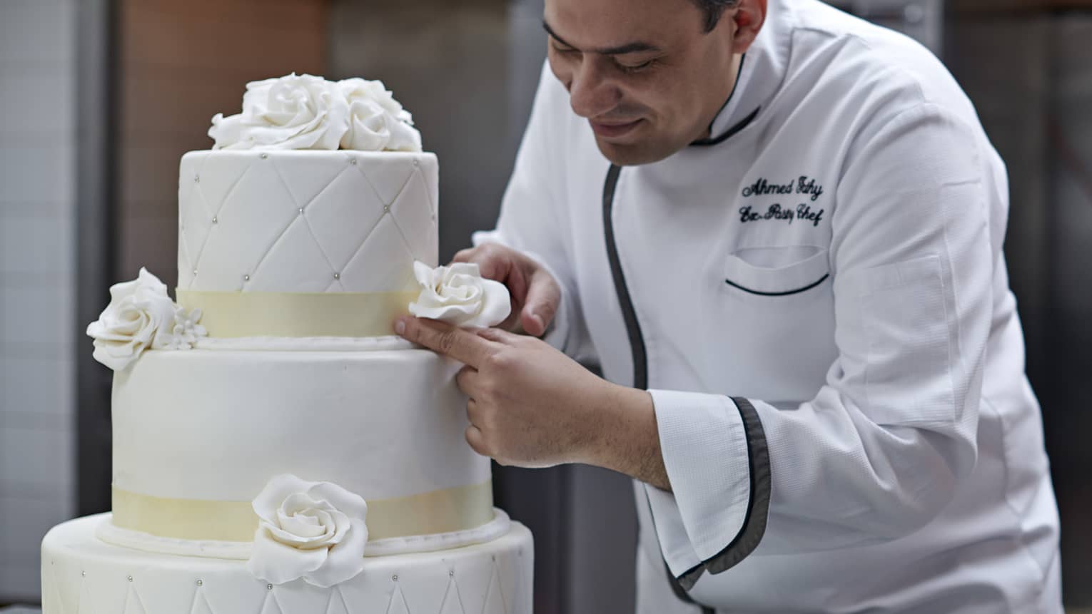Chef in white chef’s coat, leaning and putting flower detail on three-tiered white wedding cake
