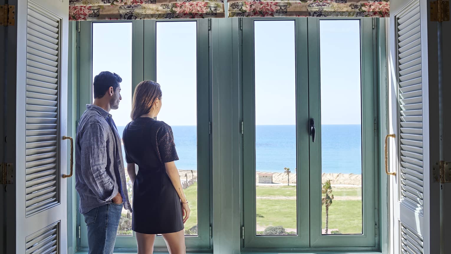 Couple stand at large rustic window overlooking Four Seasons Hotel Alexandria lawns, waterfront 