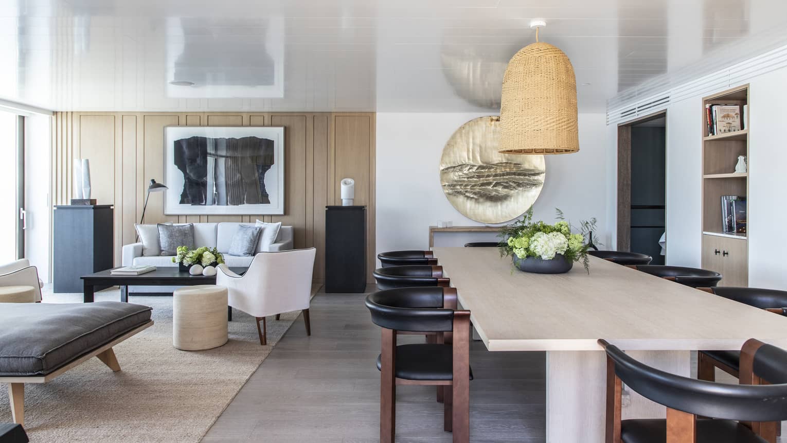 Light wood dining table, eight contemporary wood and black leather chairs, hanging wicker lamp, adjacent living room