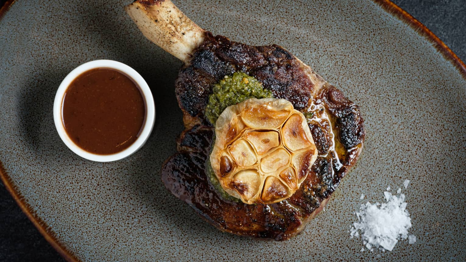 Garlic on top of a large bone-in steak on a grey plate.