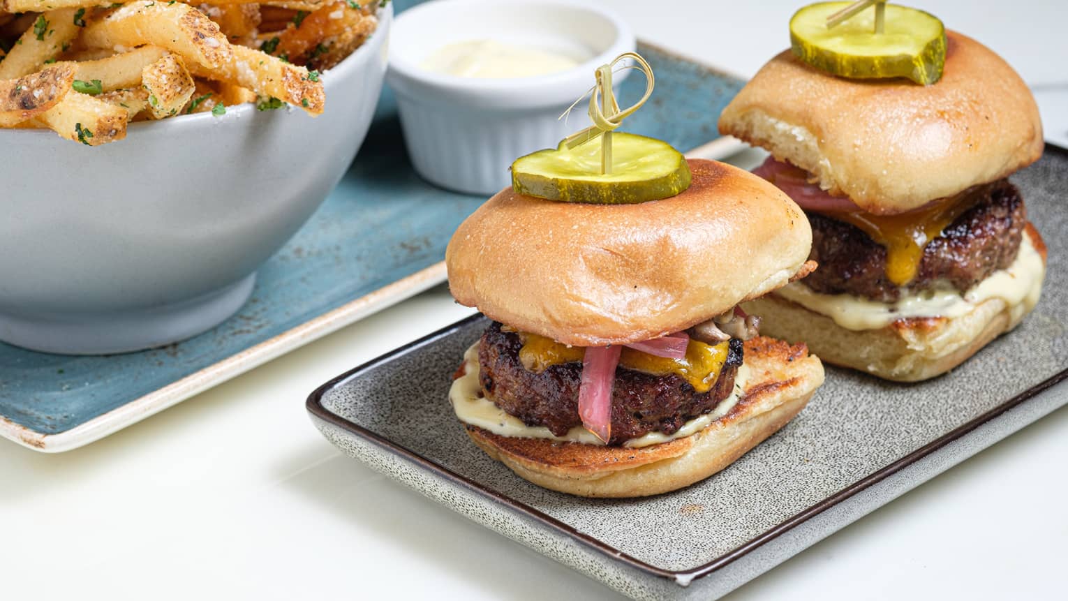 Two burgers with pickles on top of the buns and fries on rectangular plates.