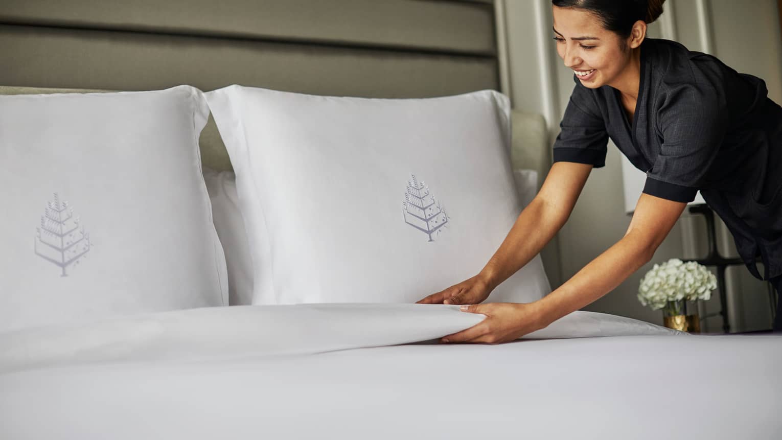 Hotel housekeeper smooths blanket under bed pillows with embroidered Four Seasons logos
