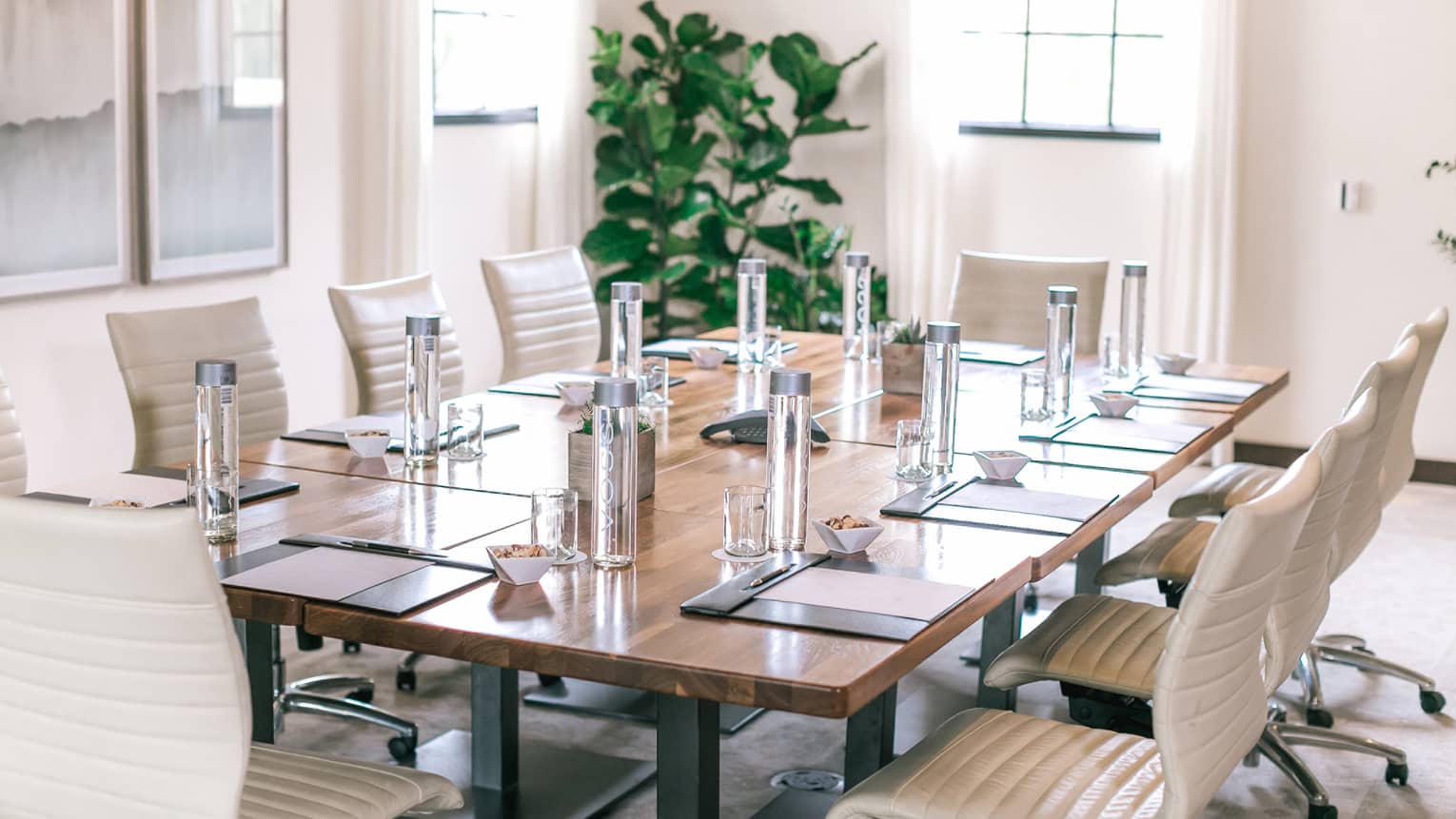 A rectangular boardroom table set with clear glass water bottles and black folders in a naturally-lit room