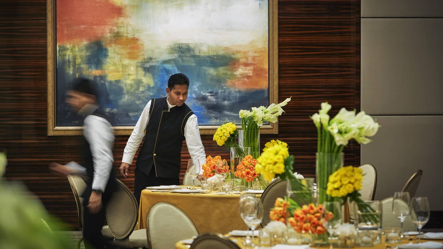 Server in black-and-white uniform sets round formal dining table with yellow linen, vases of fresh orange, yellow flowers
