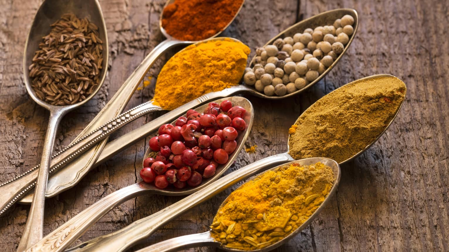 Seven silver spoons filled with various spices.