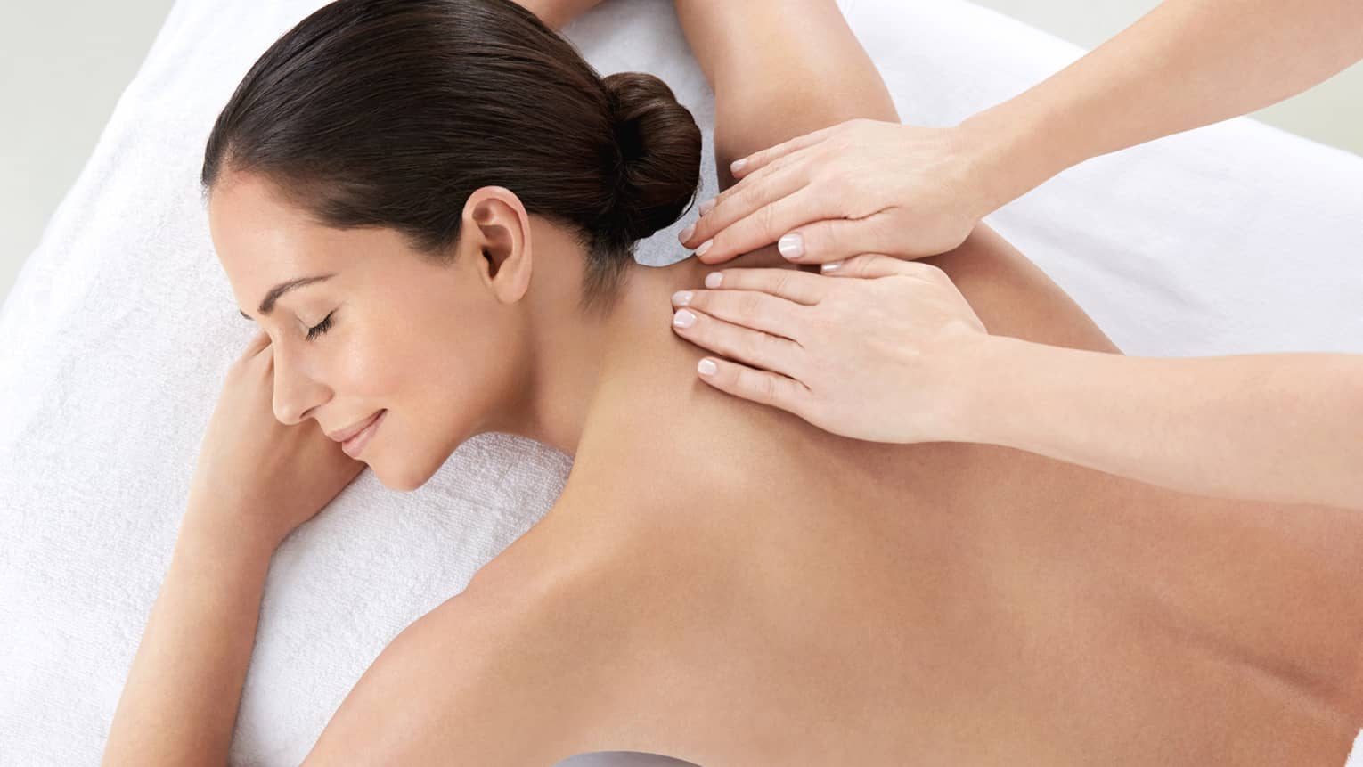 Woman smiles, lies on massage table as hands massage bare shoulders