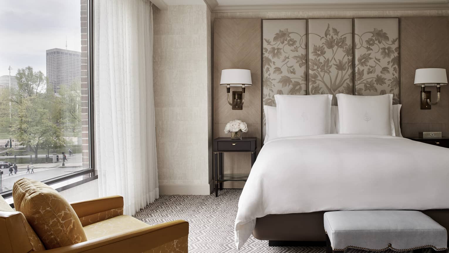 Front view of Executive Suite bed with white linens, beige fabric floral headboard, large window with white curtains, yellow armchair