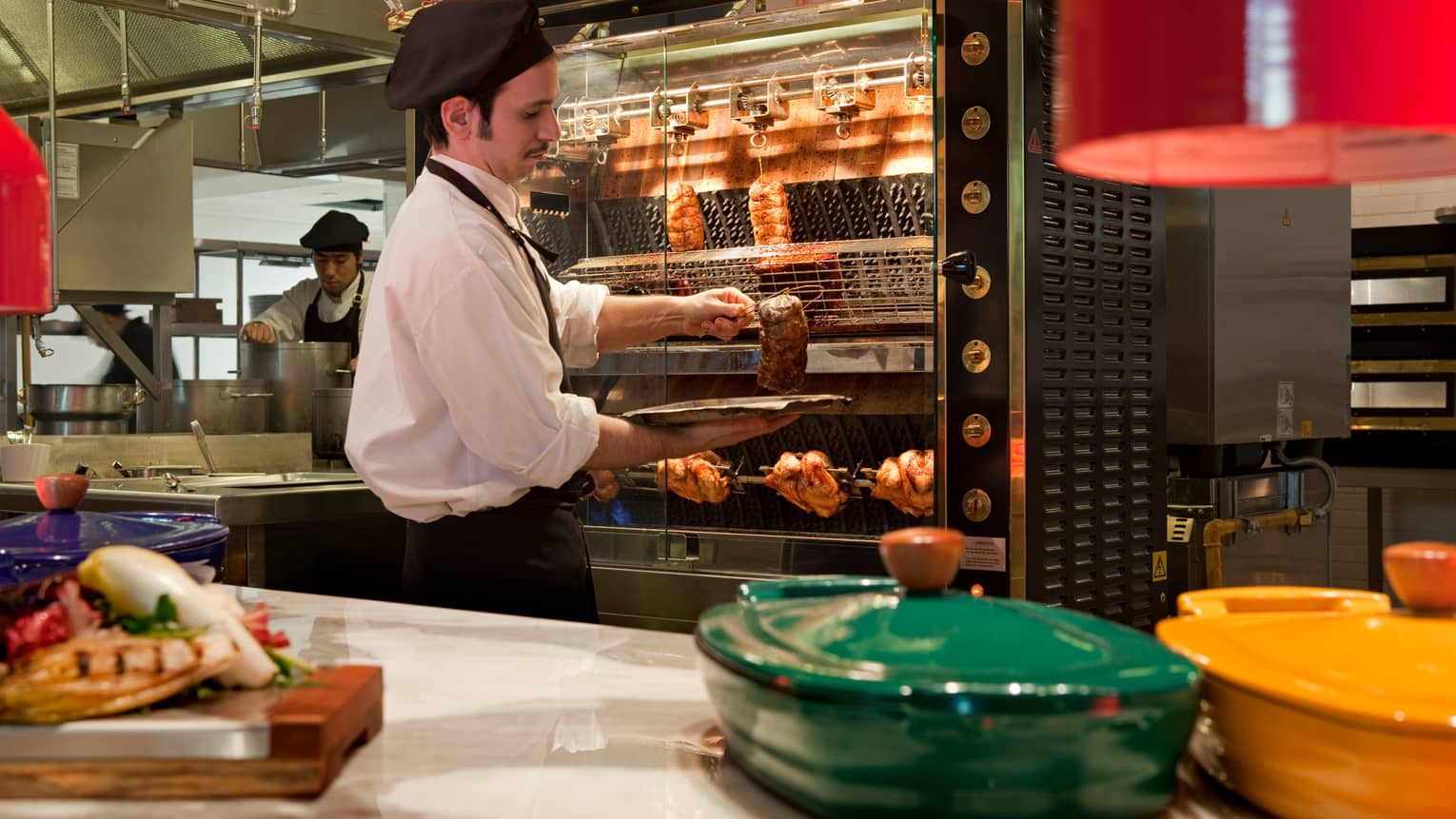 Cook takes meat out of large rotisserie in kitchen, green and yellow baking dishes with lids on counter
