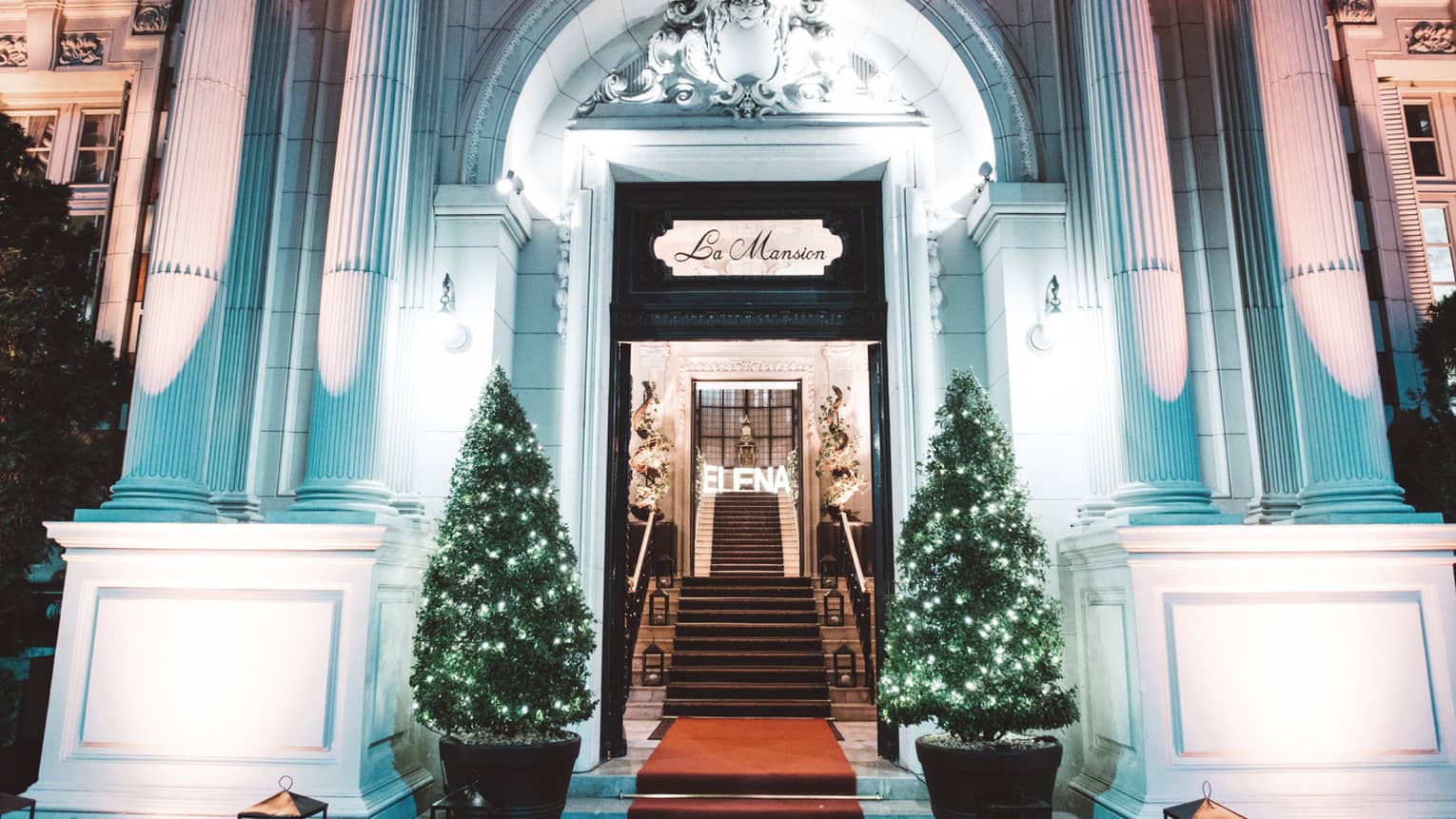 A Four Seasons Hotel Entrance is festively decorated with clear christmas lights on two small evergreen bushes