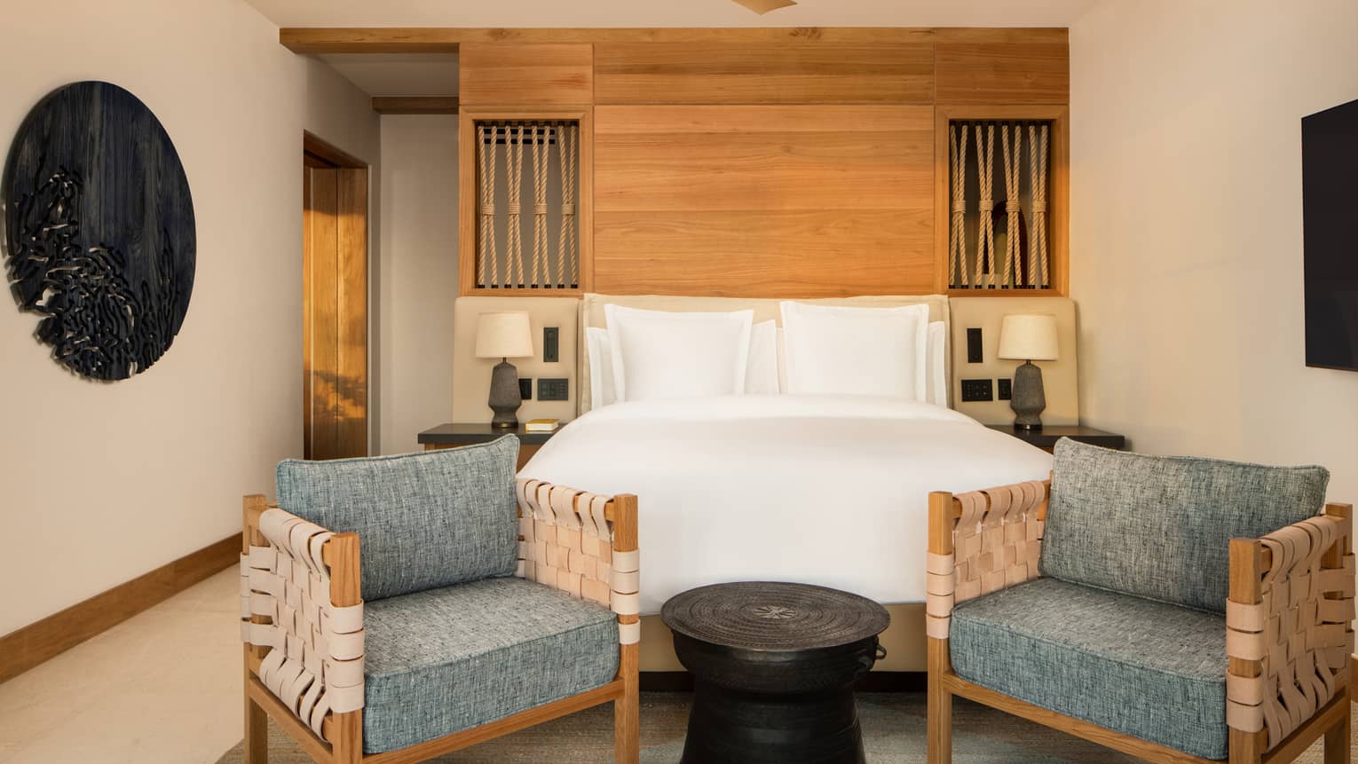 A luxury resort suite featuring a king bed, wooden accent wall, two chairs at the foot of the bed and small table