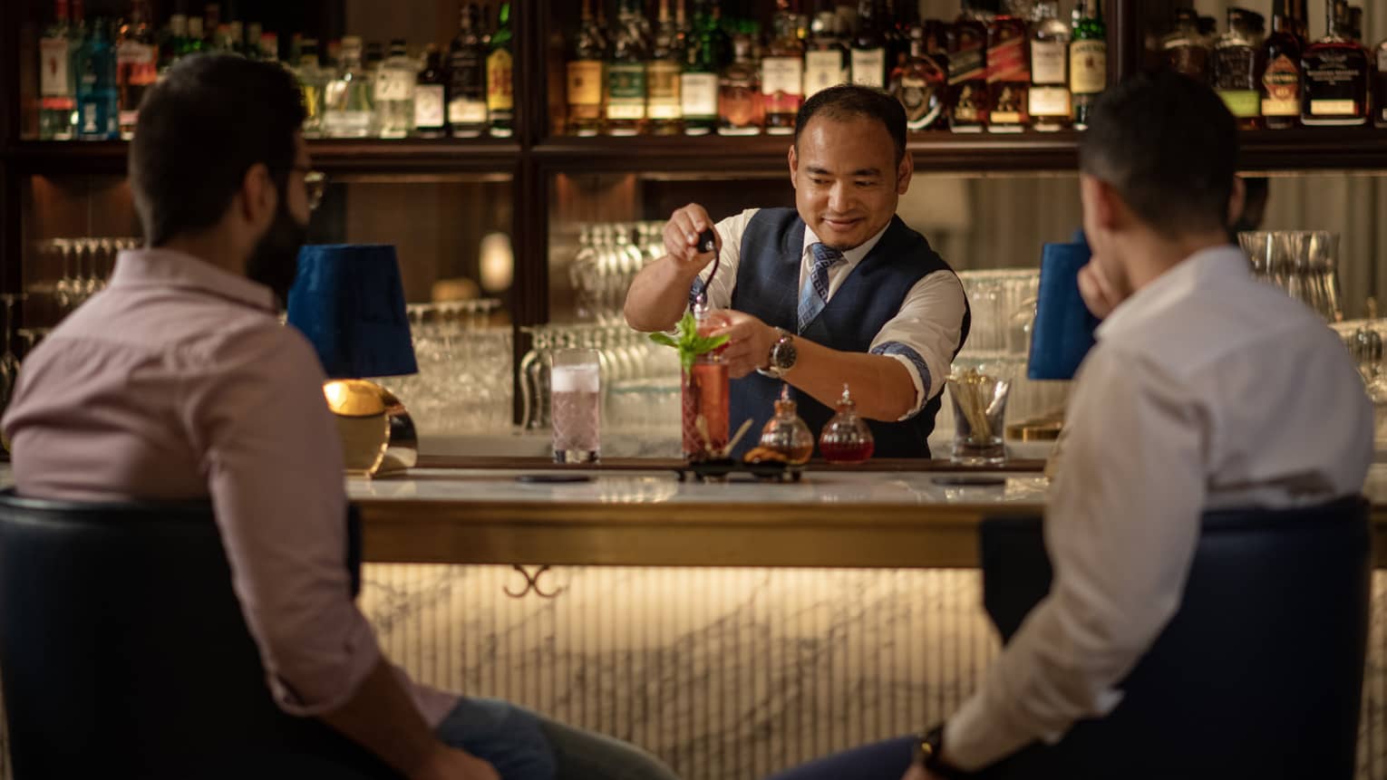 A bartender making a cocktail while two men converse at a bar.
