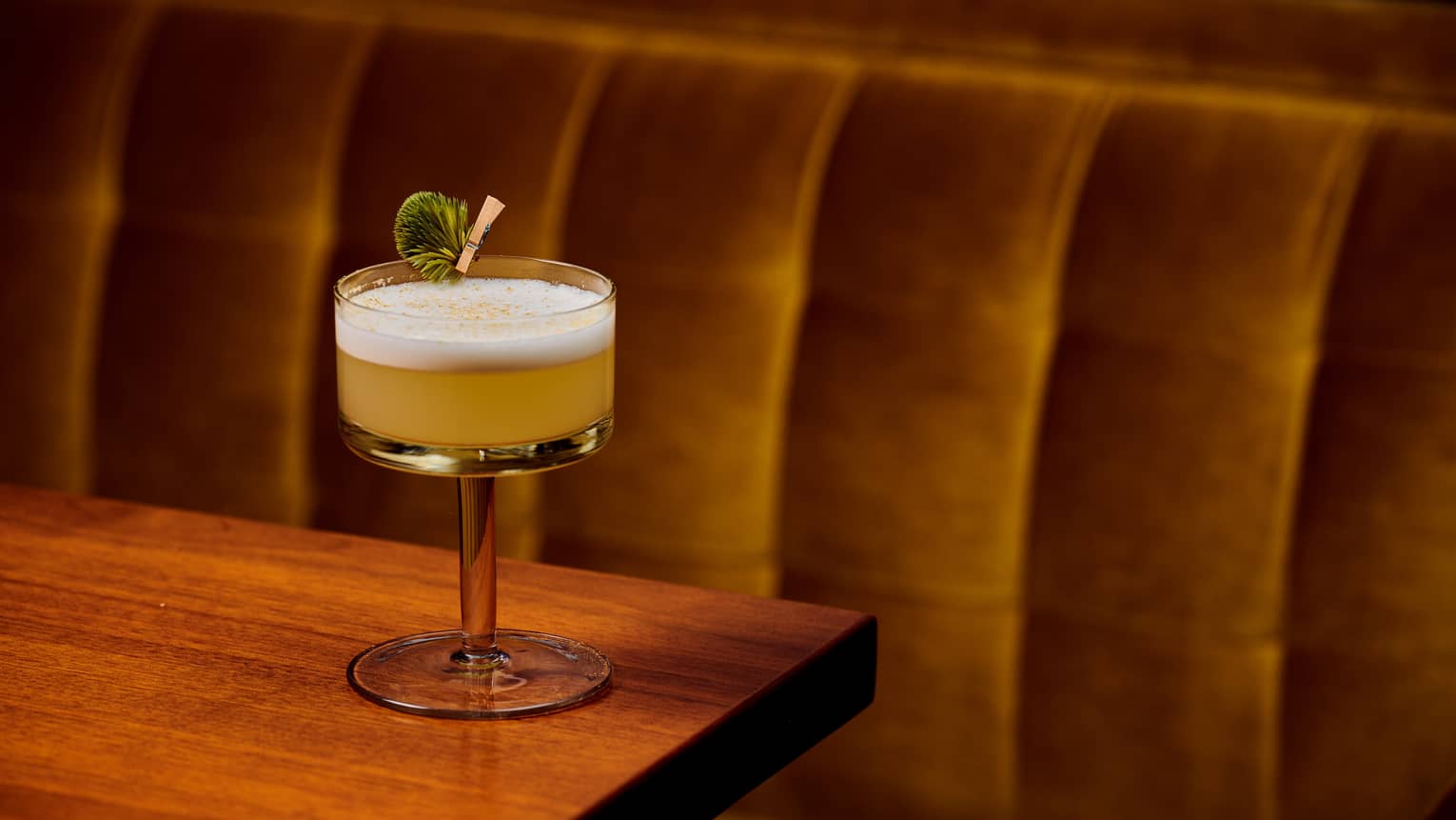 A cocktail with foam and herb garnish sits on wooden restaurant table