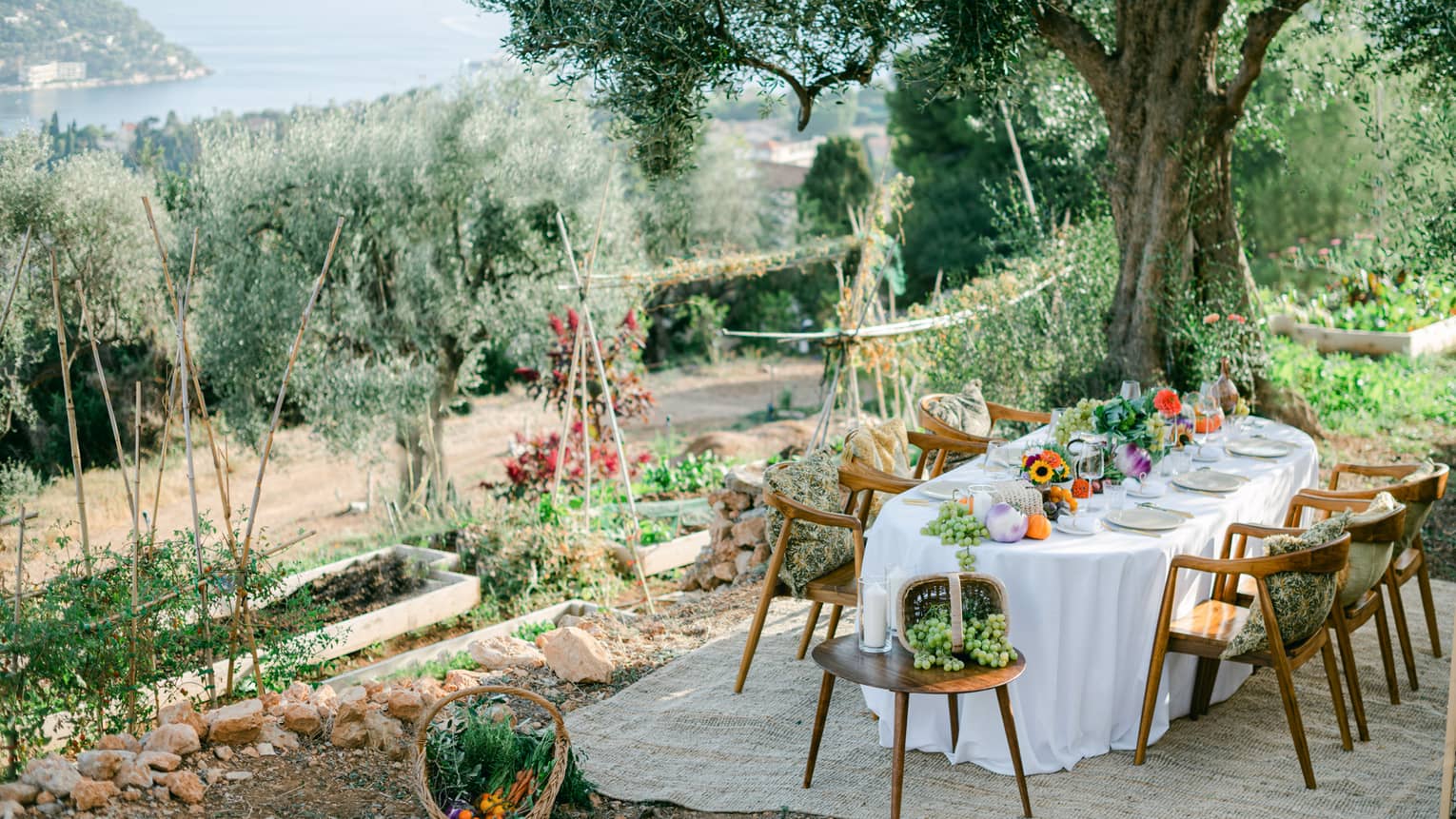 Garden dining setup with oval table draped in white and garden bounty down centre, and six chairs with pillows