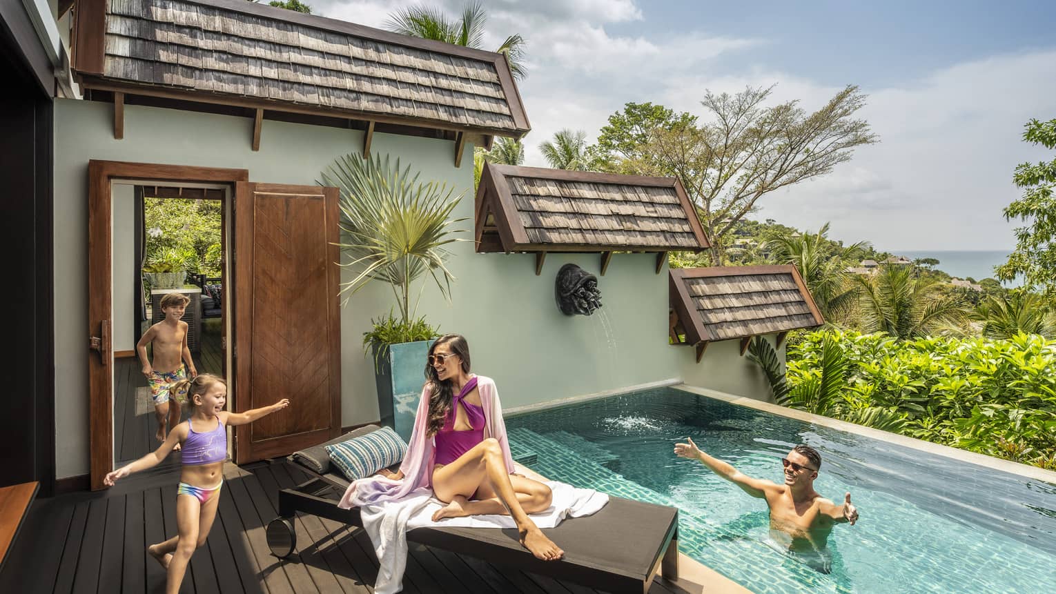 Two young children run into a small pool. Woman sits on lounge chair and man welcomes children in the pool.
