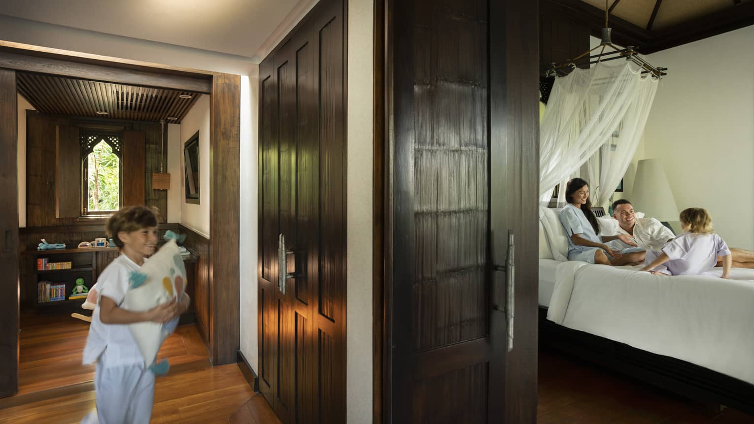 Young boy runs down a hallway to a bedroom where woman, man and small child play