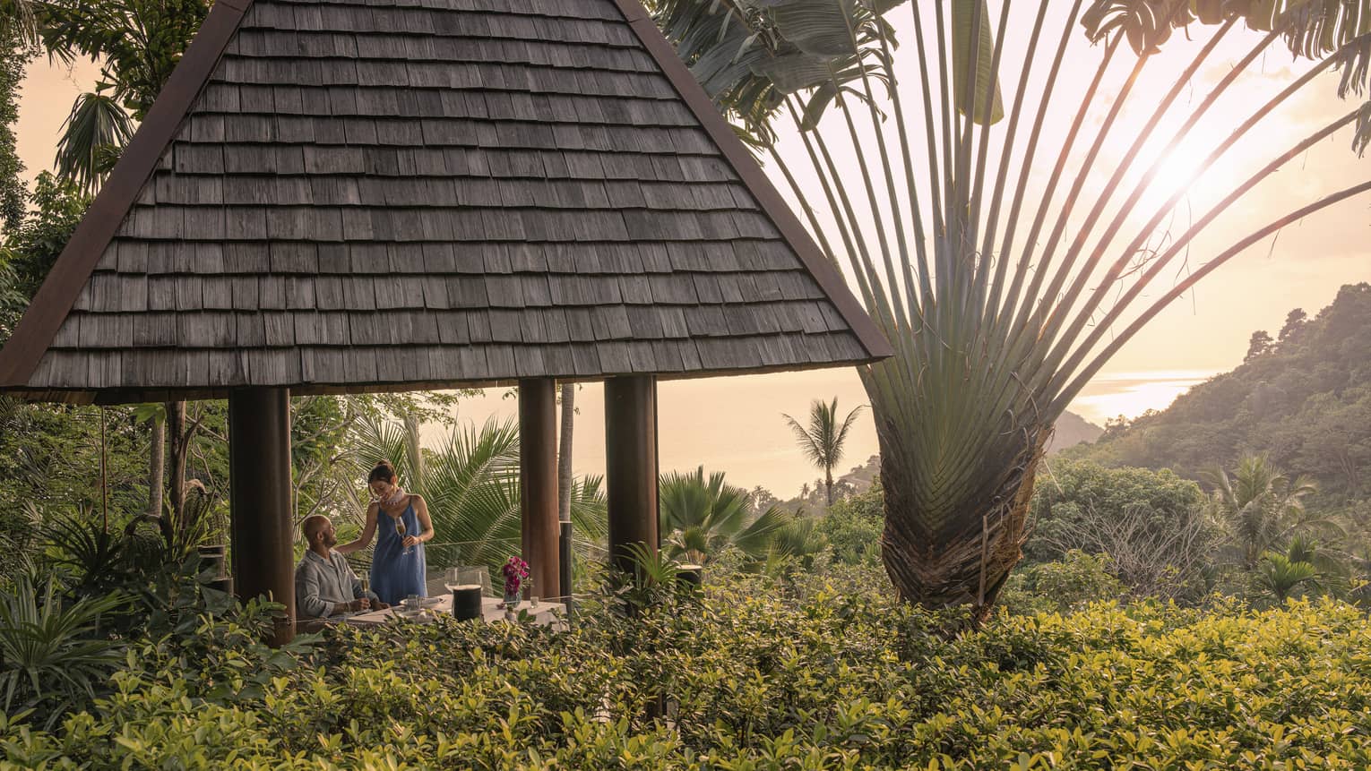 A couple with champagne in a tall hilltop pavilion with a dark, straw roof, set amidst lush forest and a giant fan palm tree.