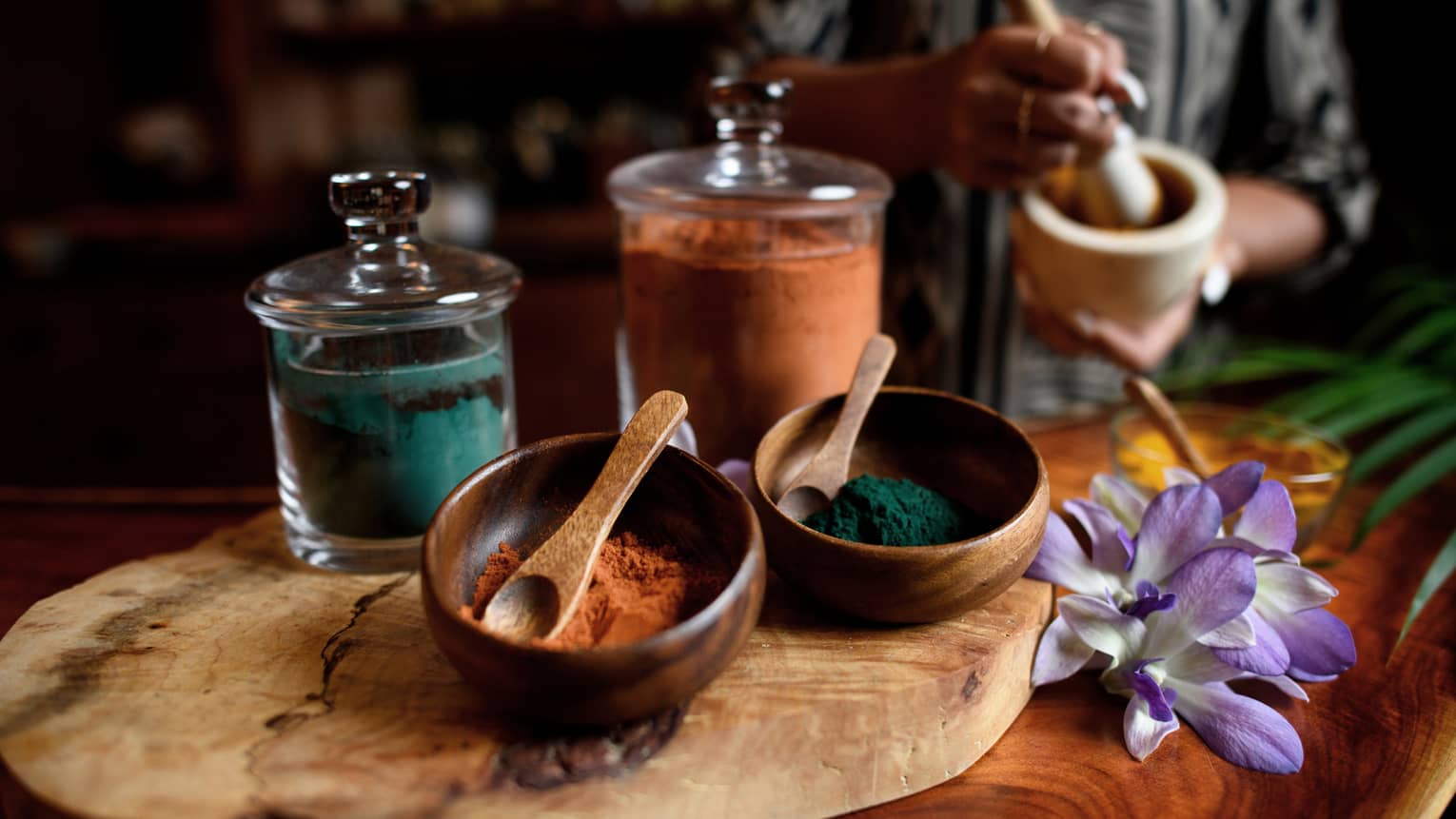 Small wood bowls and spoons beside glass jars filled with colourful powders, tropical flower